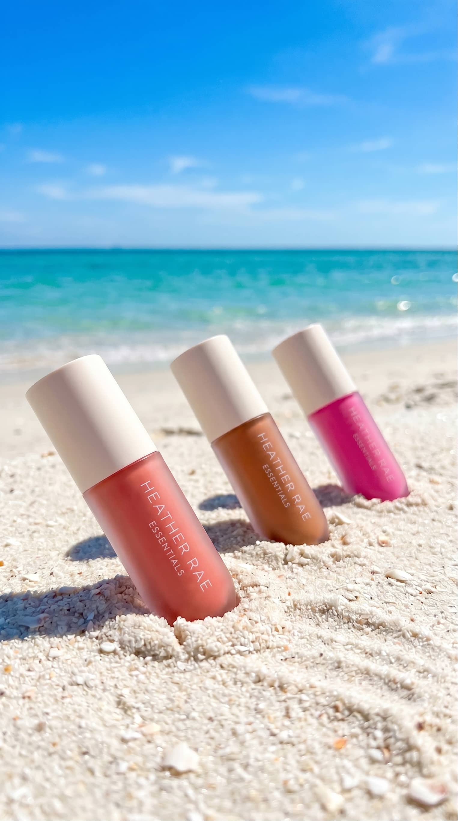 Sun-drenched coastal still life with HRE lip gloss collection resting on wet sand beside clear ocean tide refraction