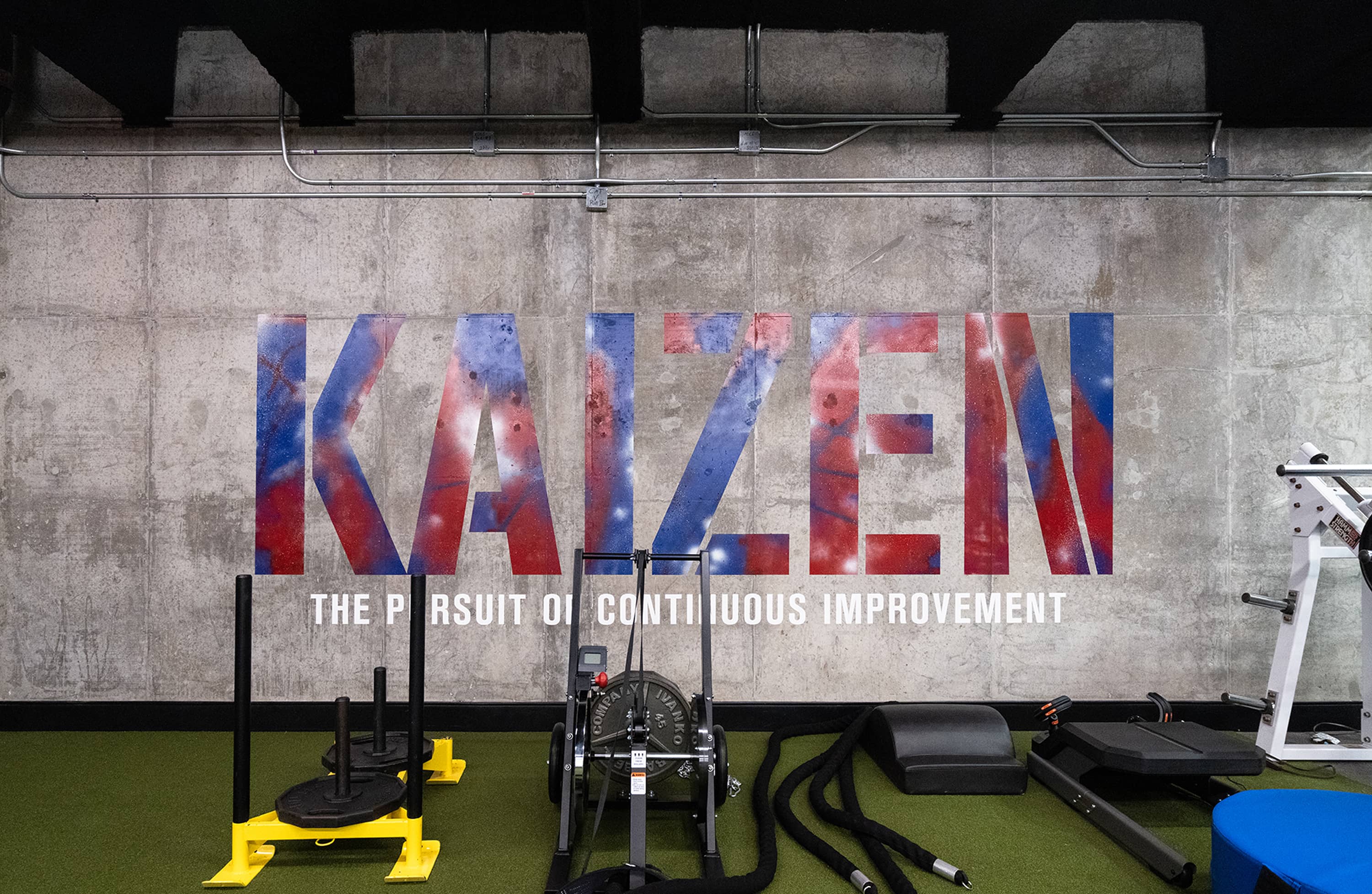 Raw concrete weight room wall stenciled with KAIZEN: The Pursuit of Continuous Improvement